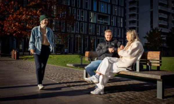 Hannah walks past Alex and Neve sitting on bench in sunshine.