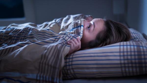 A young woman lying in bed with the covers pulled up to her face with a scared look after havin<em></em>g a nightmare